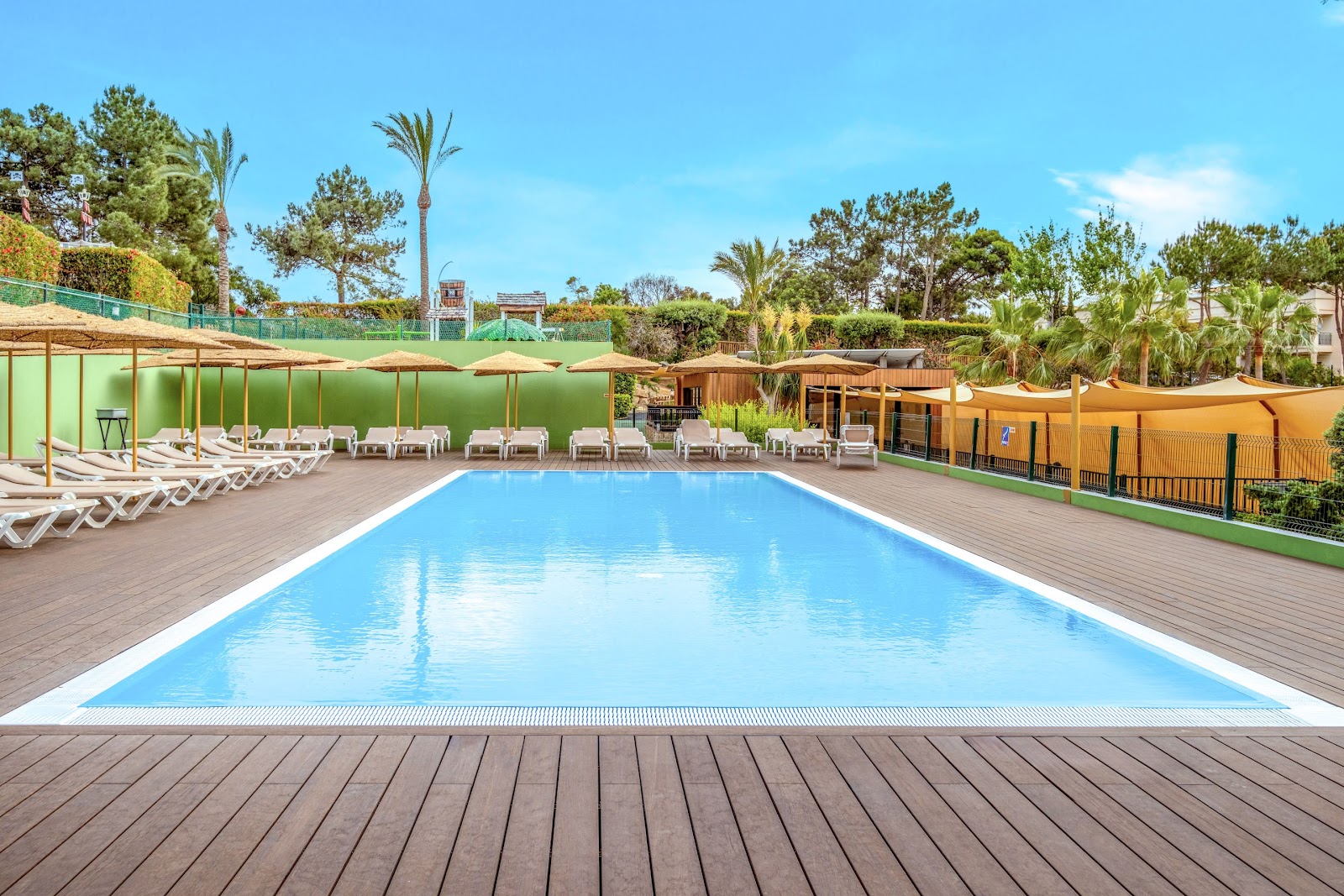 Rectangular swimming pool with wooden deck surrounded by white lounge chairs under tan umbrellas, with green wall and palm trees beyond at Hotel Guarana.