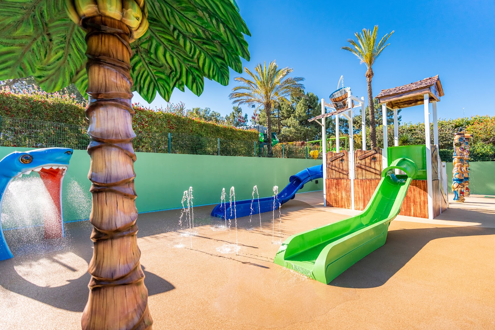 Children's water play area with green and blue slides, wooden structures, fountains, and palm trees under clear sky at Hotel Guarana.