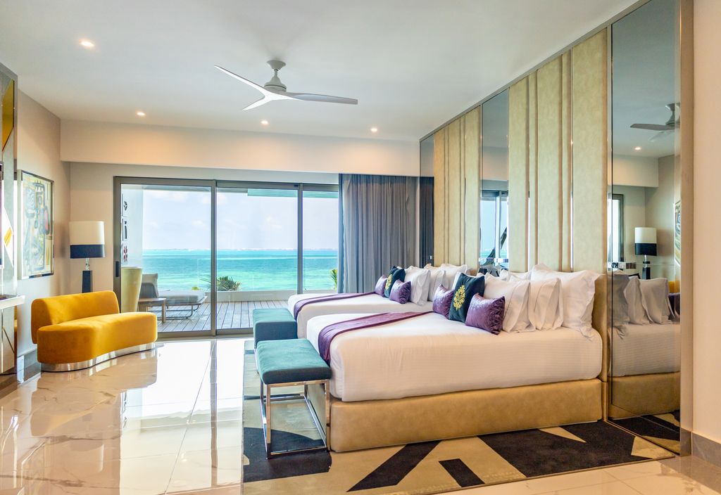 Modern oceanfront resort bedroom with two beds, purple accents, yellow armchair, and turquoise ocean view through floor-to-ceiling windows.