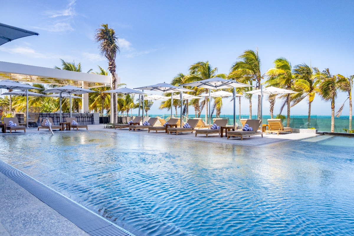 Resort pool with white umbrellas and lounge chairs overlooking the turquoise Caribbean Sea and palm trees.