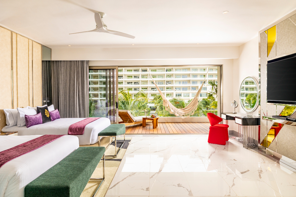 Resort suite with two beds, balcony overlooking tropical gardens and multi-story buildings, hammock on wooden deck, flat-screen TV, and red chair.