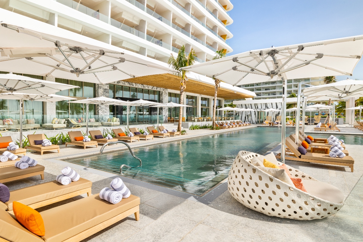 Resort pool deck with loungers, white umbrellas, rolled towels, and a modern white high-rise building under clear blue sky.