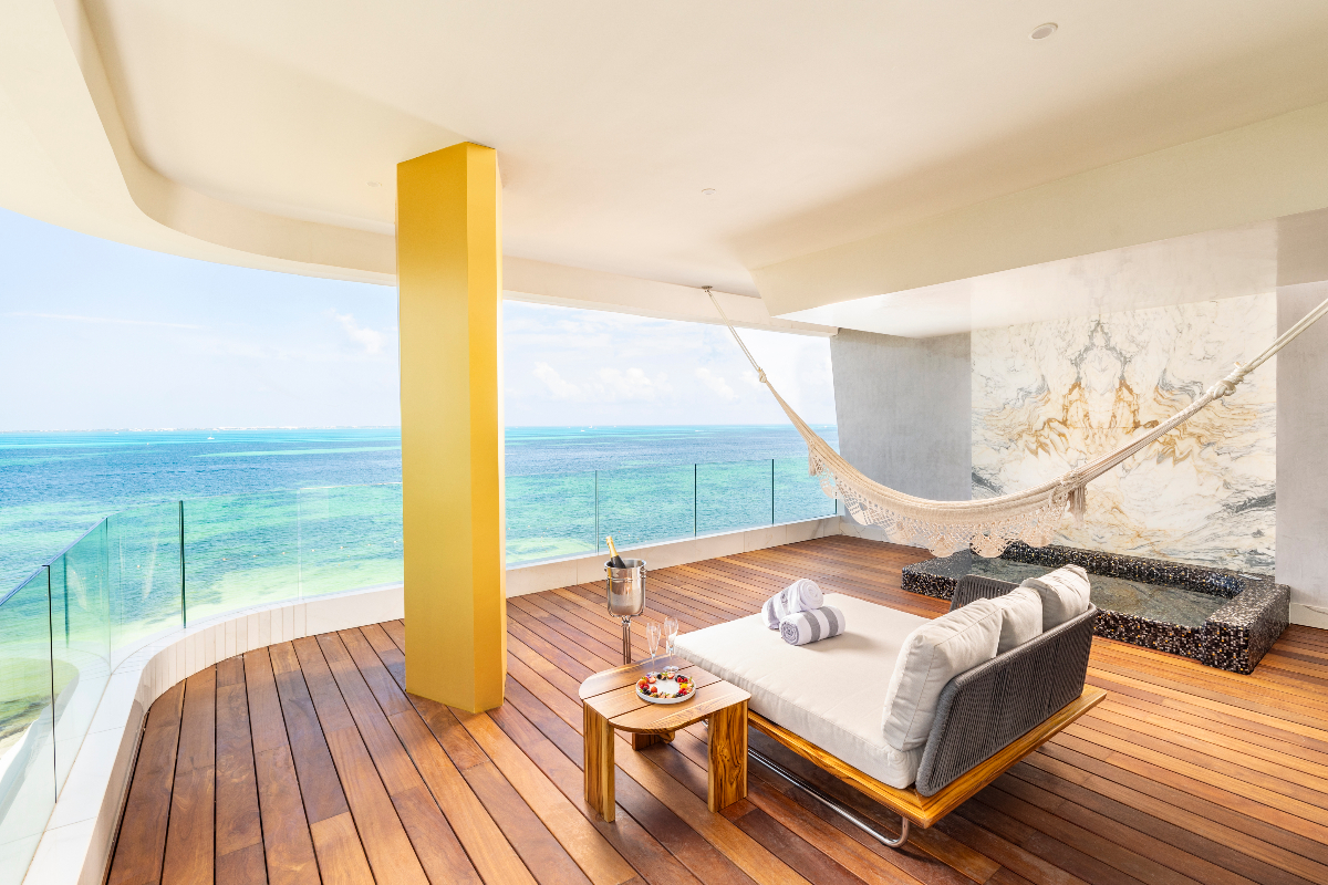 Open-air oceanview terrace with wooden deck, gray lounge seating, yellow support column, hammock, and turquoise Caribbean waters beyond glass railing.
