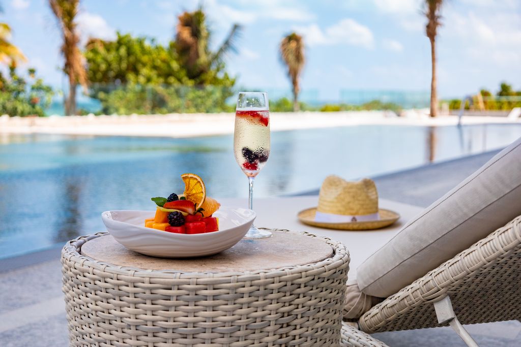 Sparkling fruit cocktail and fresh fruit bowl on a wicker table by the pool at Hotel Mousai Cancun, with palm trees and ocean view beyond.