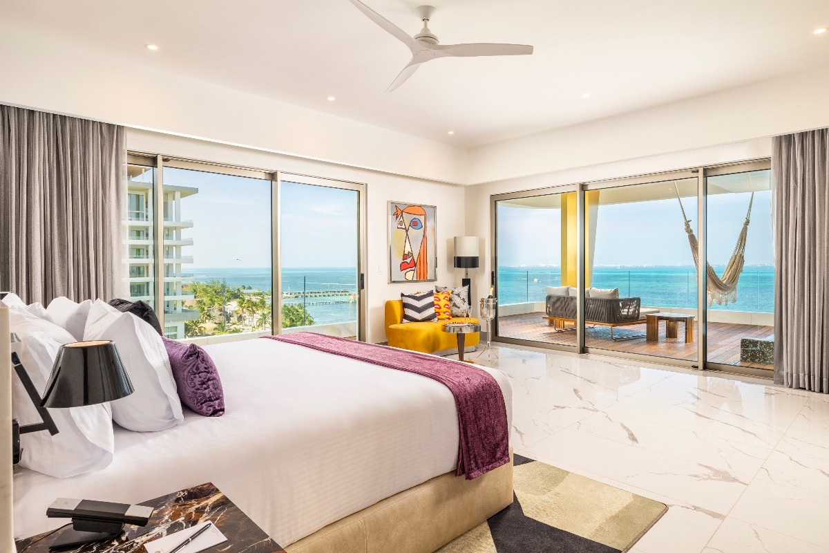 Modern hotel suite with ocean-view windows, white bed with purple accent, sitting area, and balcony overlooking Caribbean Sea.