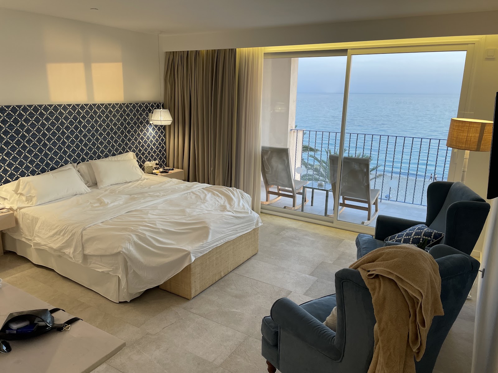 Oceanfront resort bedroom with king bed, patterned headboard, and seating area overlooking the Mediterranean Sea from a balcony.