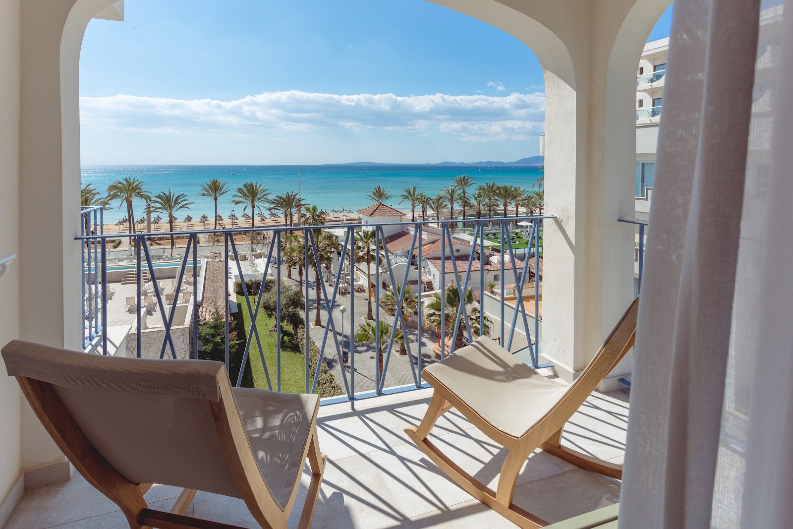 Two lounge chairs on a resort balcony overlook palm-lined beach, turquoise sea, and distant mountains under blue sky.