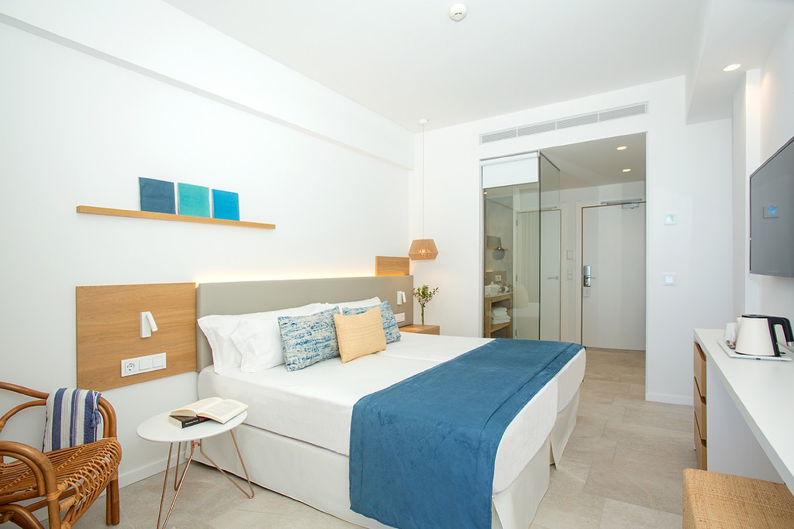 Bright modern hotel room with white walls, queen bed in blue and white linens, wood nightstands, glass shower enclosure, and wicker chair.