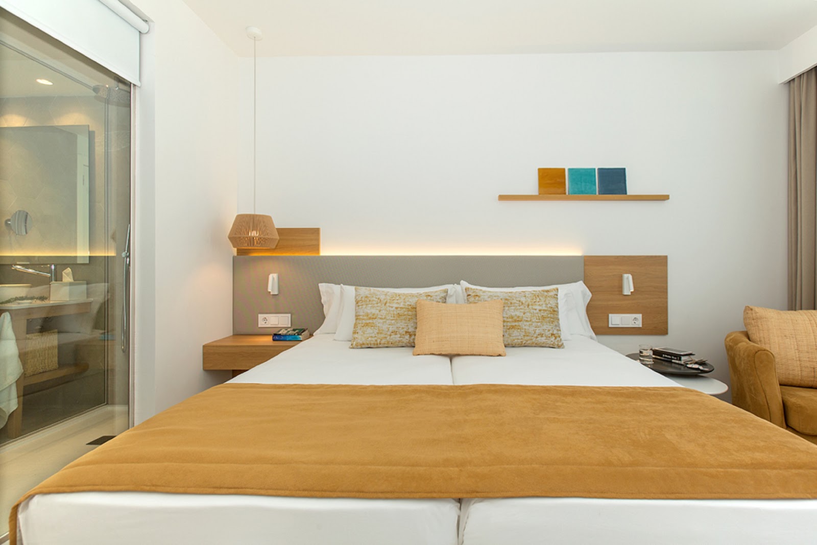Modern resort bedroom with queen bed, wood headboard with shelving, glass shower enclosure, and neutral furnishings in warm tones.