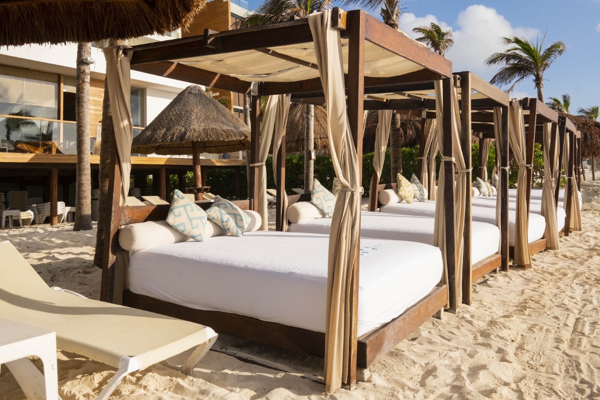 Row of wooden cabana beds with white mattresses and draped curtains on a sandy beach near palm trees and thatched structures.
