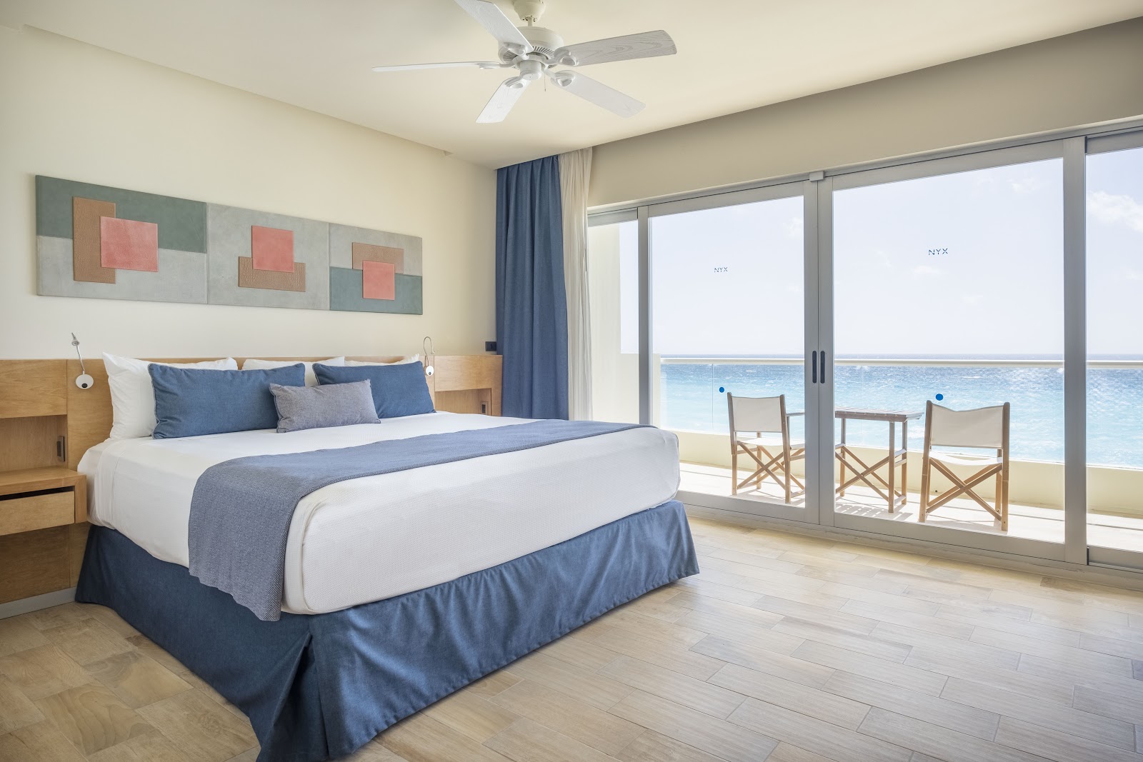Hotel NYX Cancun beachfront room with queen bed, blue bedding, and glass doors opening to ocean view and beach chairs.