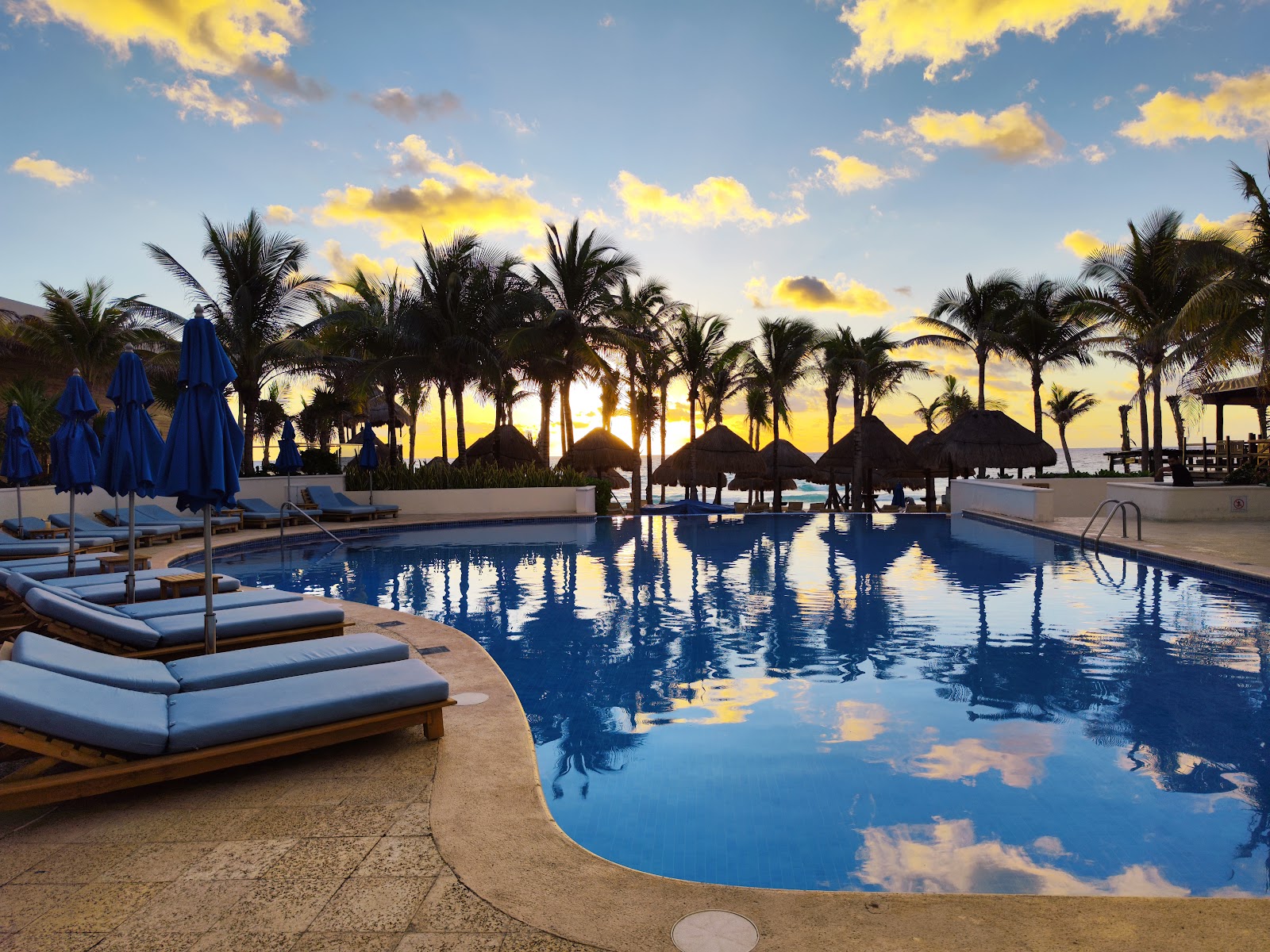 Resort pool at sunset with blue umbrellas, lounge chairs, palm trees, and thatched palapas reflected in the water.