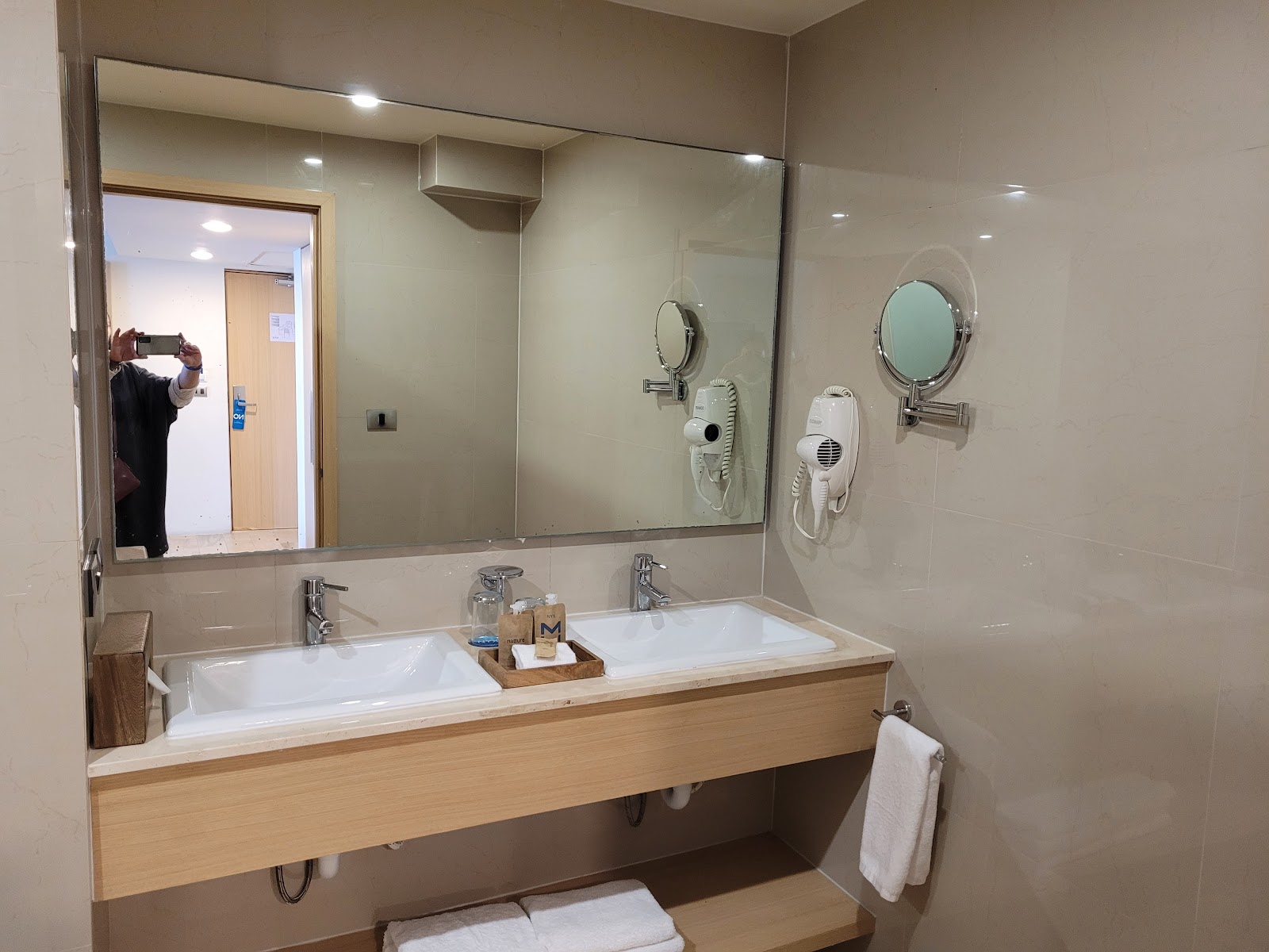 Modern bathroom with dual white sinks, large mirror, wall-mounted hair dryers, and wooden vanity at Hotel NYX Cancun.