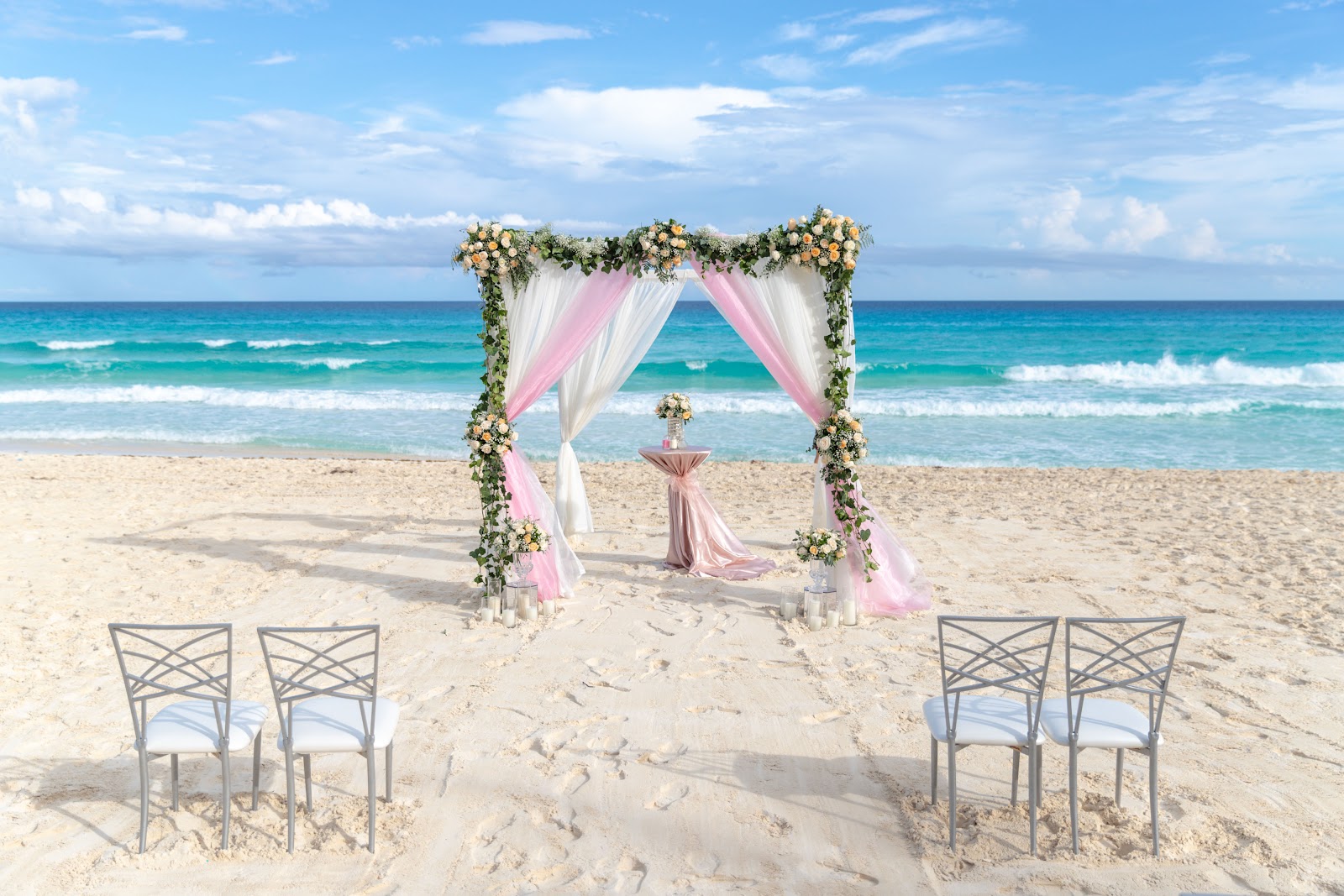 Beach wedding setup at Hotel NYX Cancun with pink and white draped arch decorated with yellow flowers, altar table, and guest seating facing turquoise ocean.