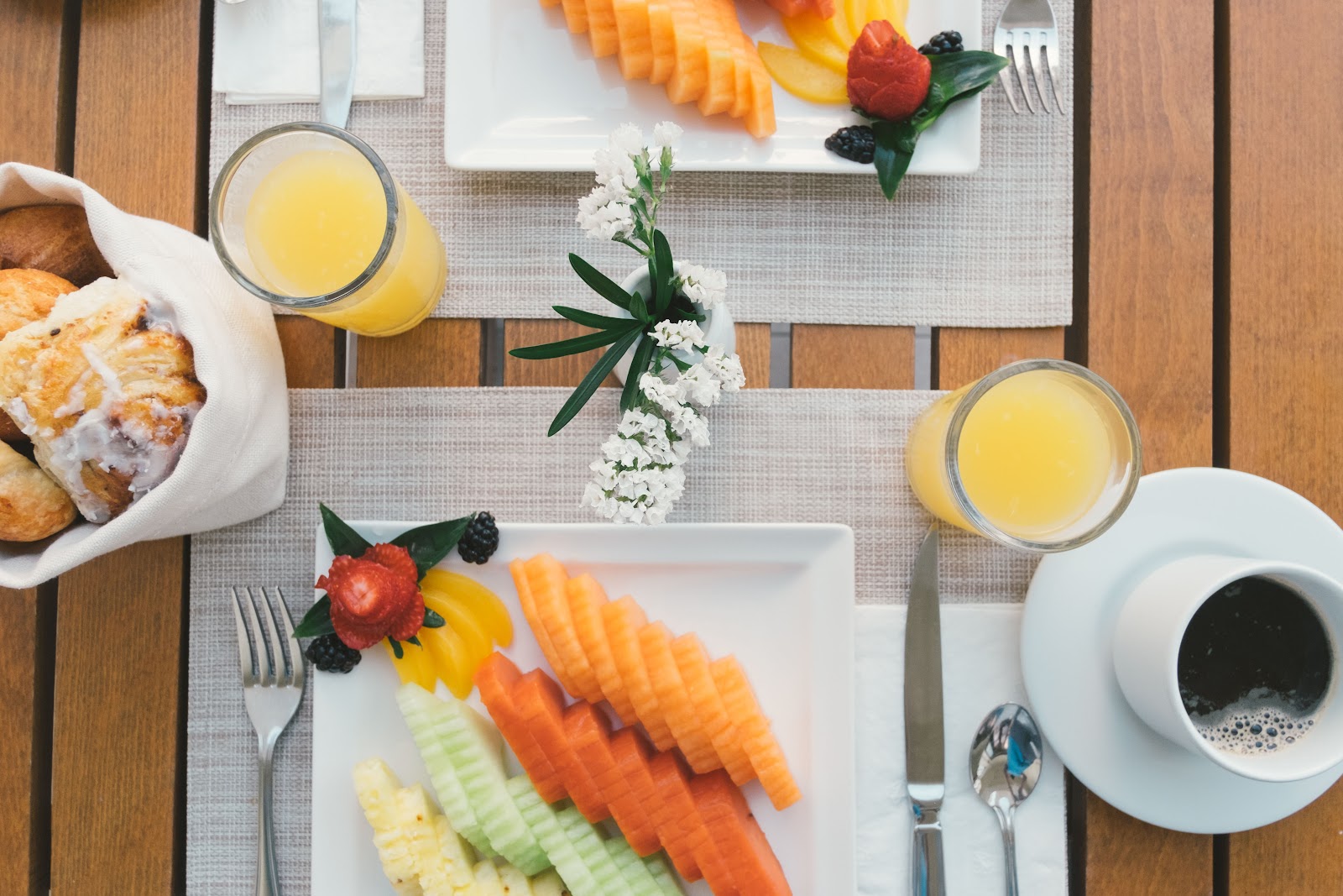 Overhead view of a breakfast table with fresh fruit, pastries, orange juice, and coffee on a wooden deck.
