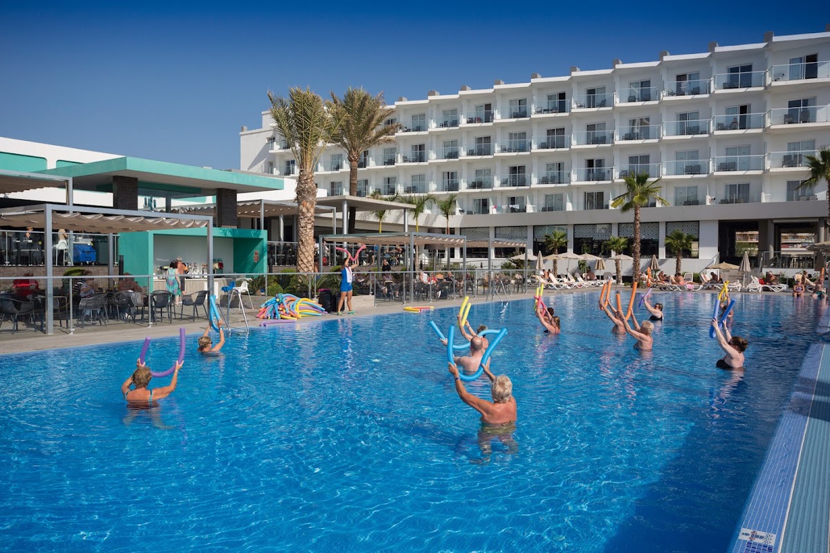 Guests participate in a pool exercise class at the Hotel Riu Costa del Sol, with a white multi-story building and turquoise bar structure in the background.