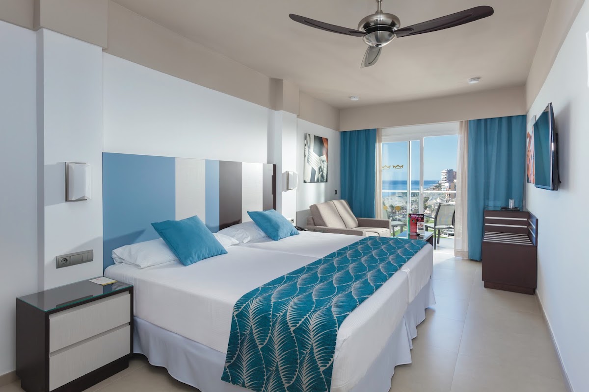 Resort room with two double beds, blue headboard, teal curtains, ceiling fan, and ocean view from balcony doors.