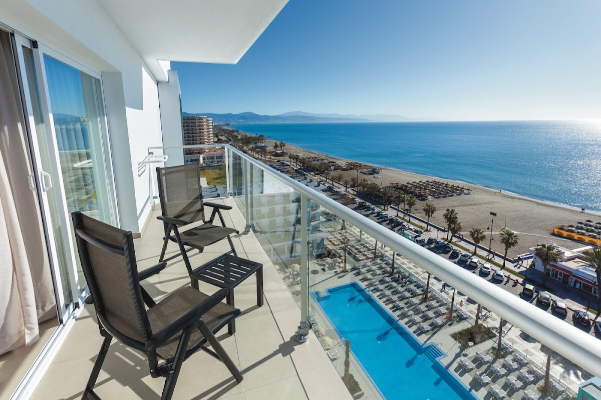 Balcony with two black chairs overlooking a blue pool, beach, and Mediterranean Sea at Hotel Riu Costa del Sol.