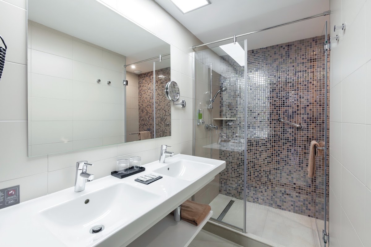 Modern bathroom with white countertop, dual sinks, large mirror, and walk-in shower with brown mosaic tile walls at Hotel Riu Costa del Sol.