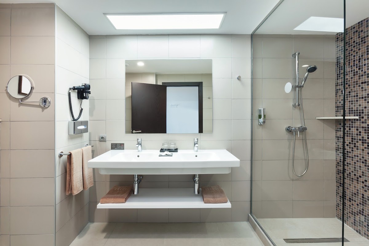Modern bathroom with dual white sink, large mirror, rainfall showerhead, and brown mosaic tile accent wall at Hotel Riu Costa del Sol.