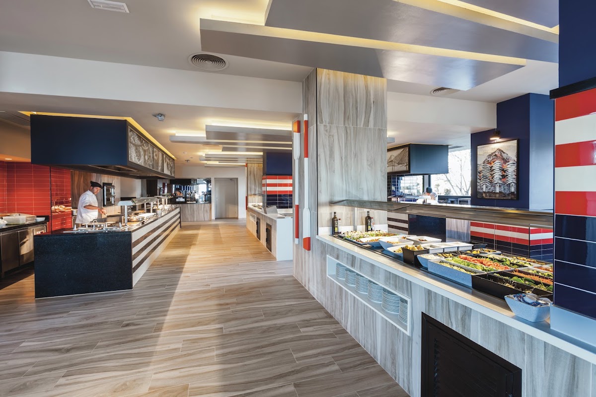 Modern dining hall at Hotel Riu Costa del Sol with red and blue striped walls, multiple food stations, and wood-look flooring.