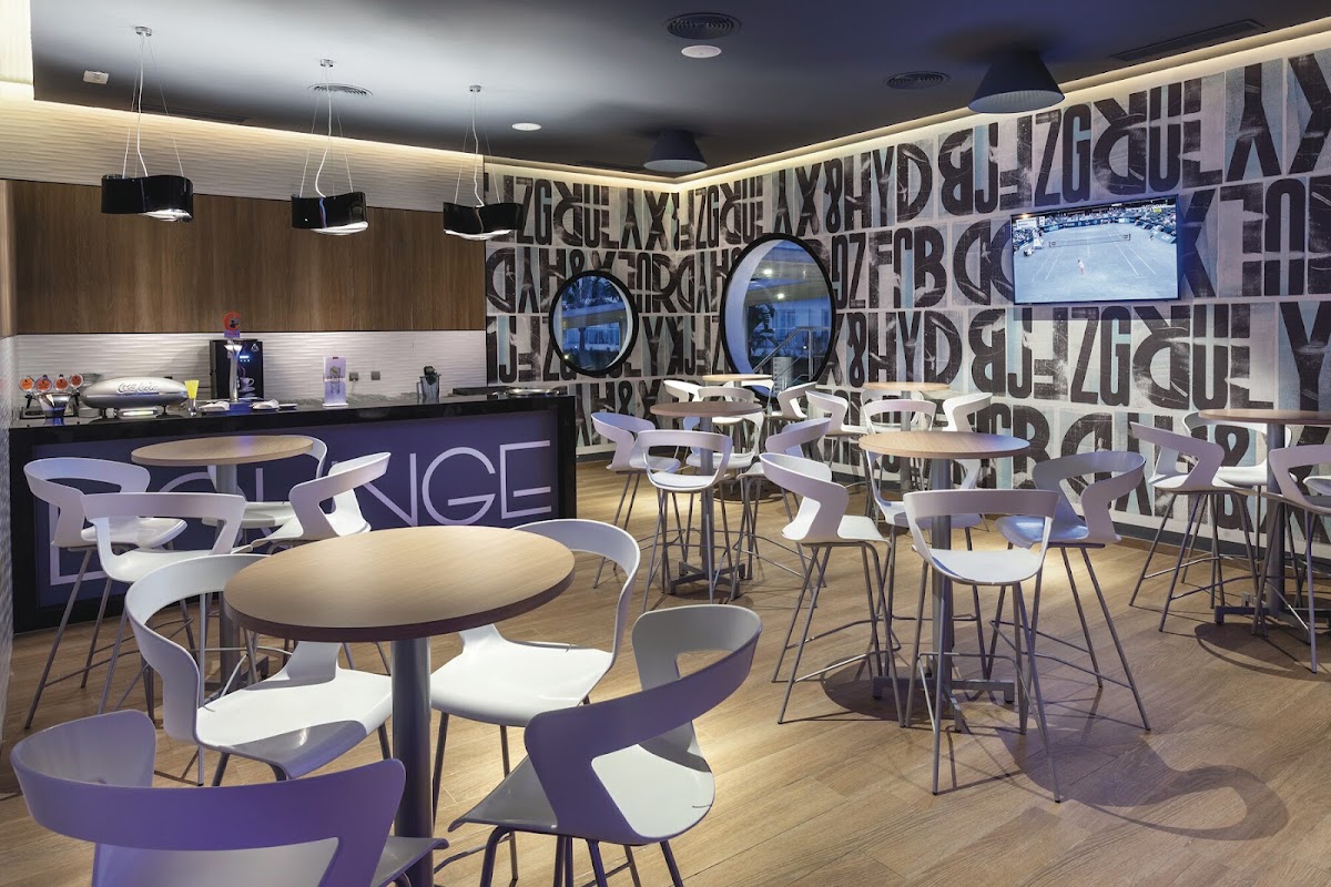 Modern lounge bar with purple and white chairs around round tables, a service counter, and TV screens on a graphic accent wall.