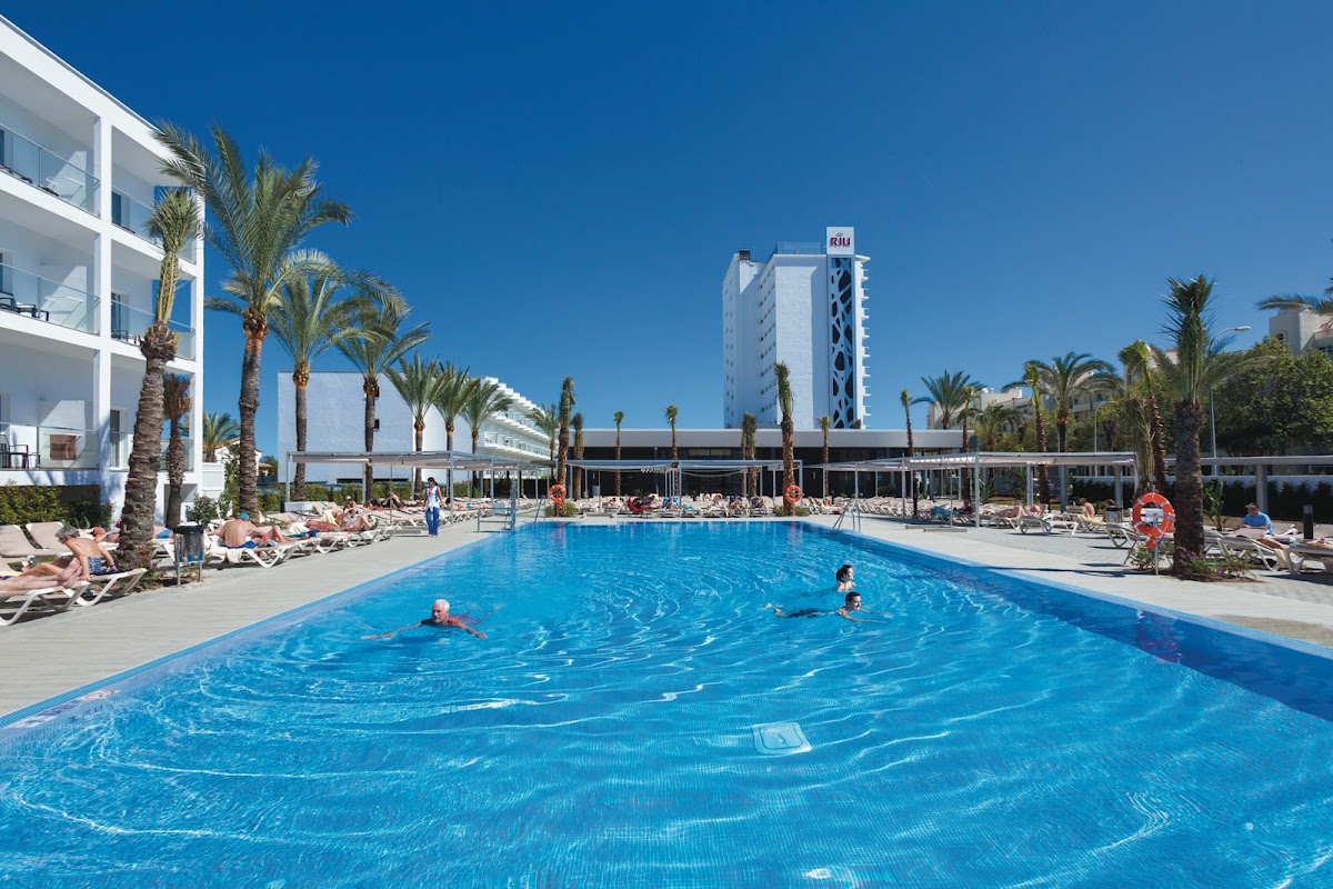 Blue pool with swimmers surrounded by palm trees, loungers, and white hotel buildings under clear sky at Hotel Riu Costa del Sol.