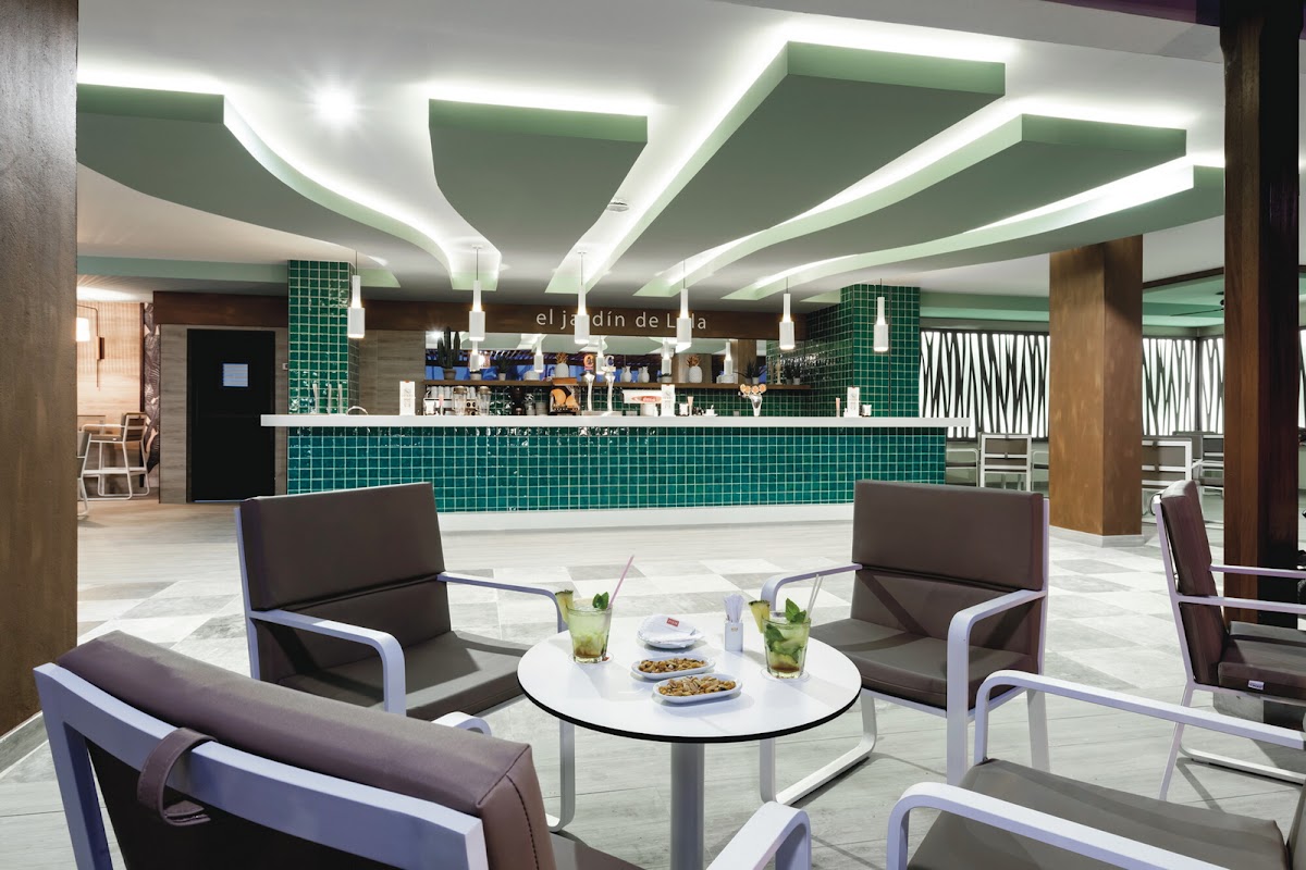 Modern lounge at Hotel Riu Costa del Sol with gray armchairs around a white table, green tile bar backdrop, and curved ceiling with LED lighting.