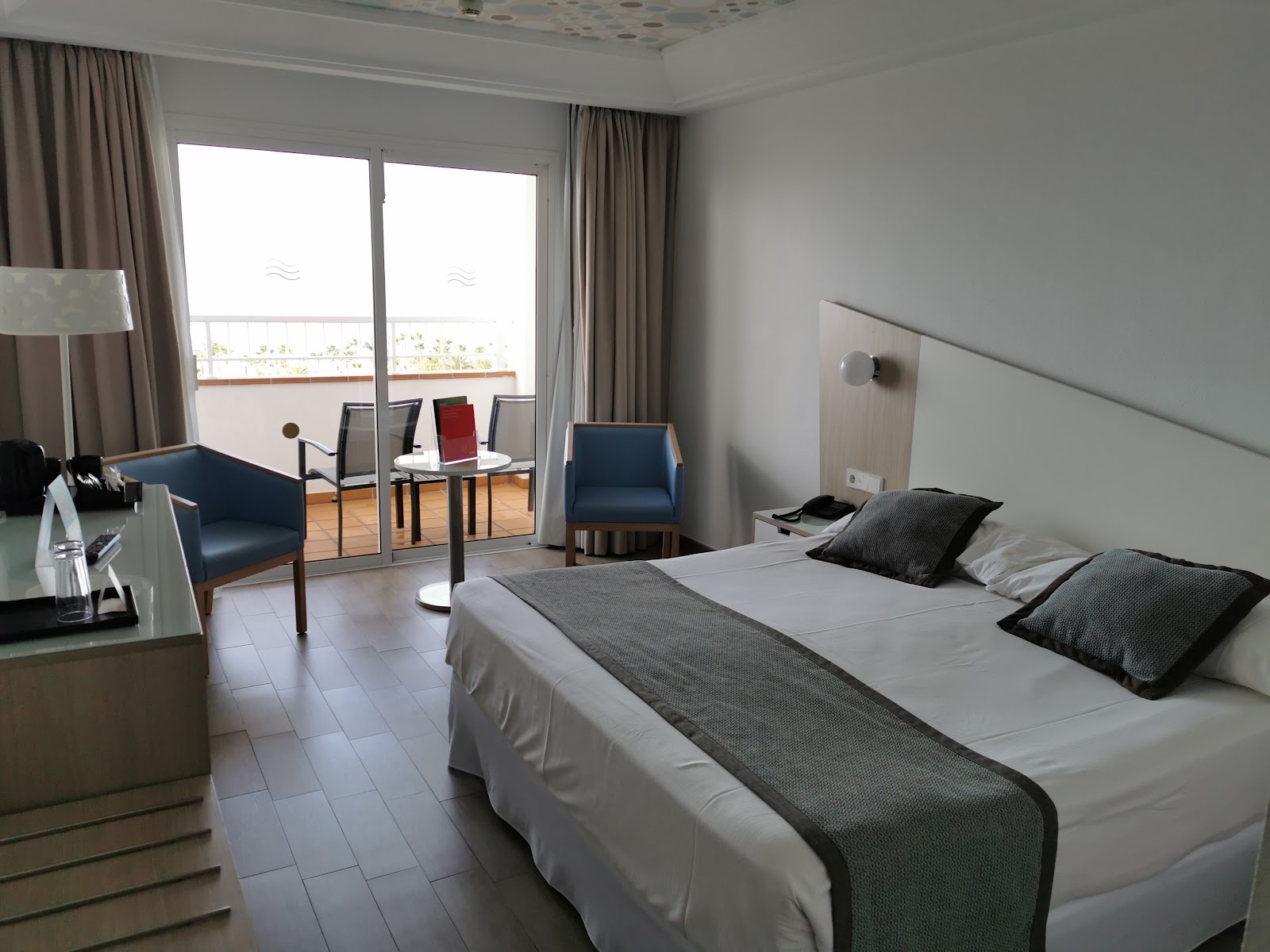 Resort guest room with double bed, sitting area with blue chairs, and balcony overlooking the coast at Hotel Riu Gran Canaria.