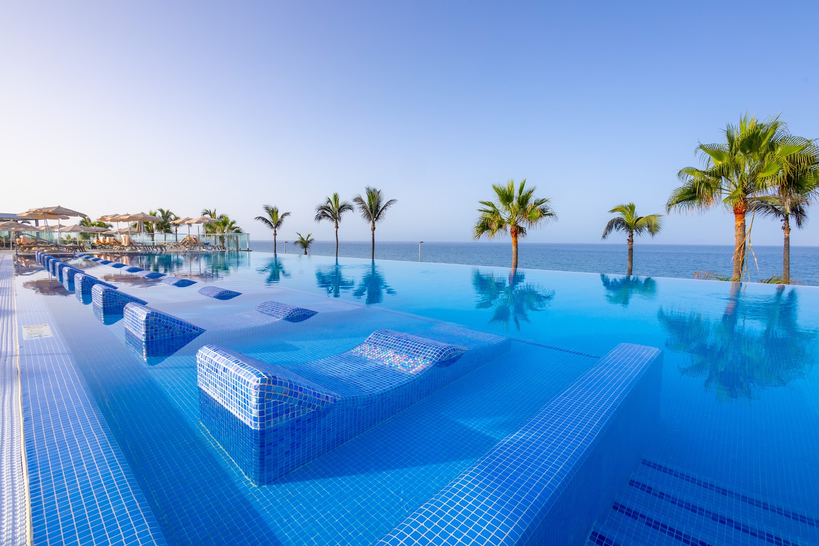 Infinity pool with sunbeds and palm trees overlooking the ocean under clear blue sky at Hotel Riu Gran Canaria.