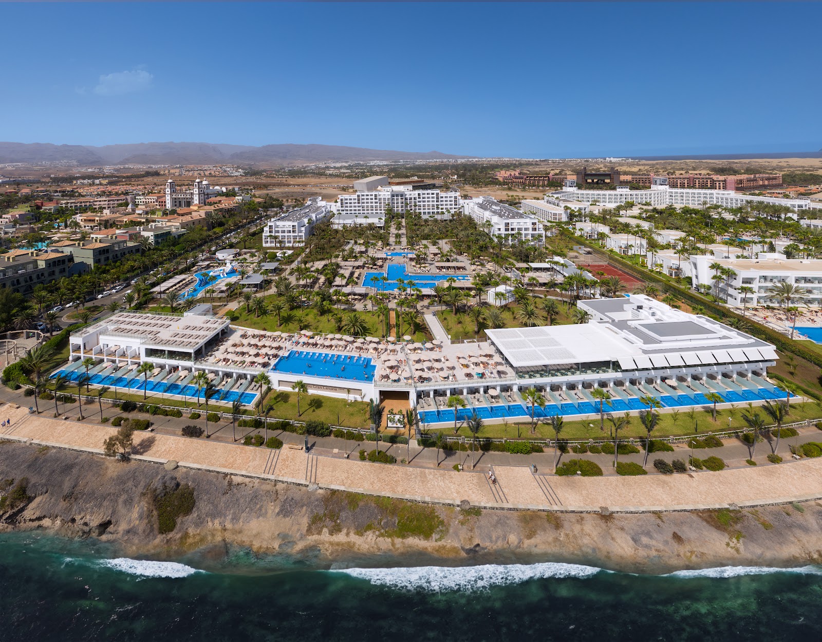 Aerial view of Hotel Riu Gran Canaria beachfront resort with pools, palm trees, sandy beach, and coastal cliffs along the Atlantic Ocean.