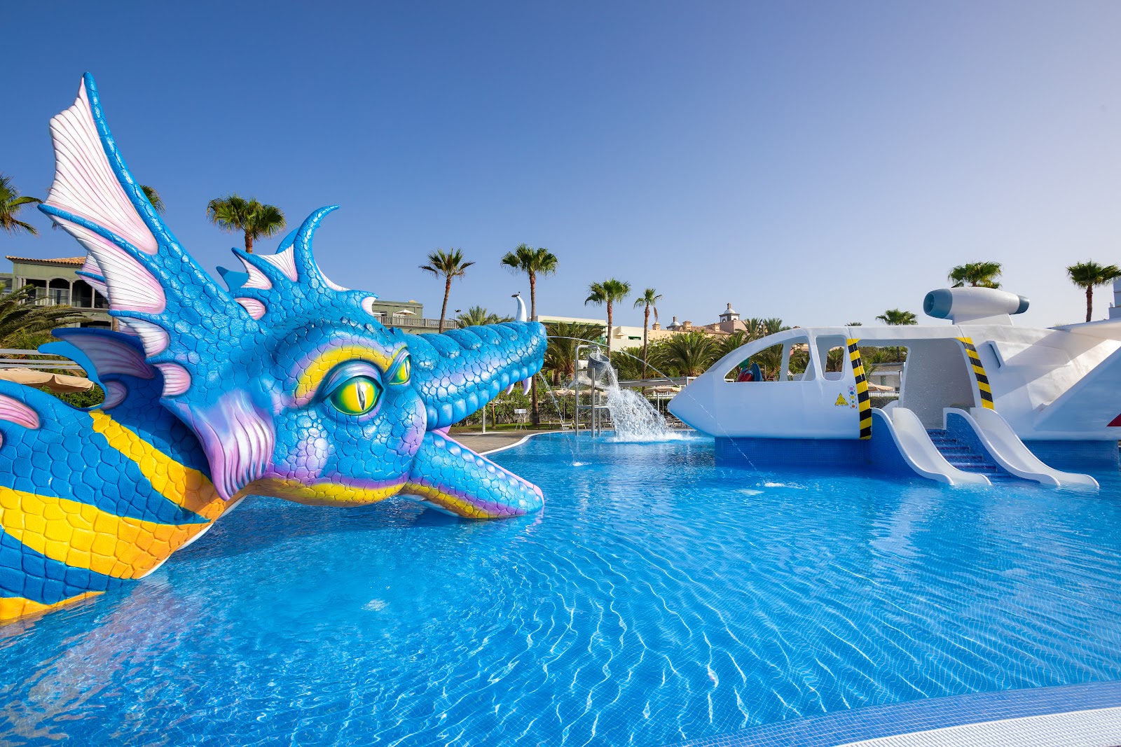 Colorful dragon and airplane water slides overlook a bright blue pool at Hotel Riu Gran Canaria, with palm trees and resort buildings in the background.
