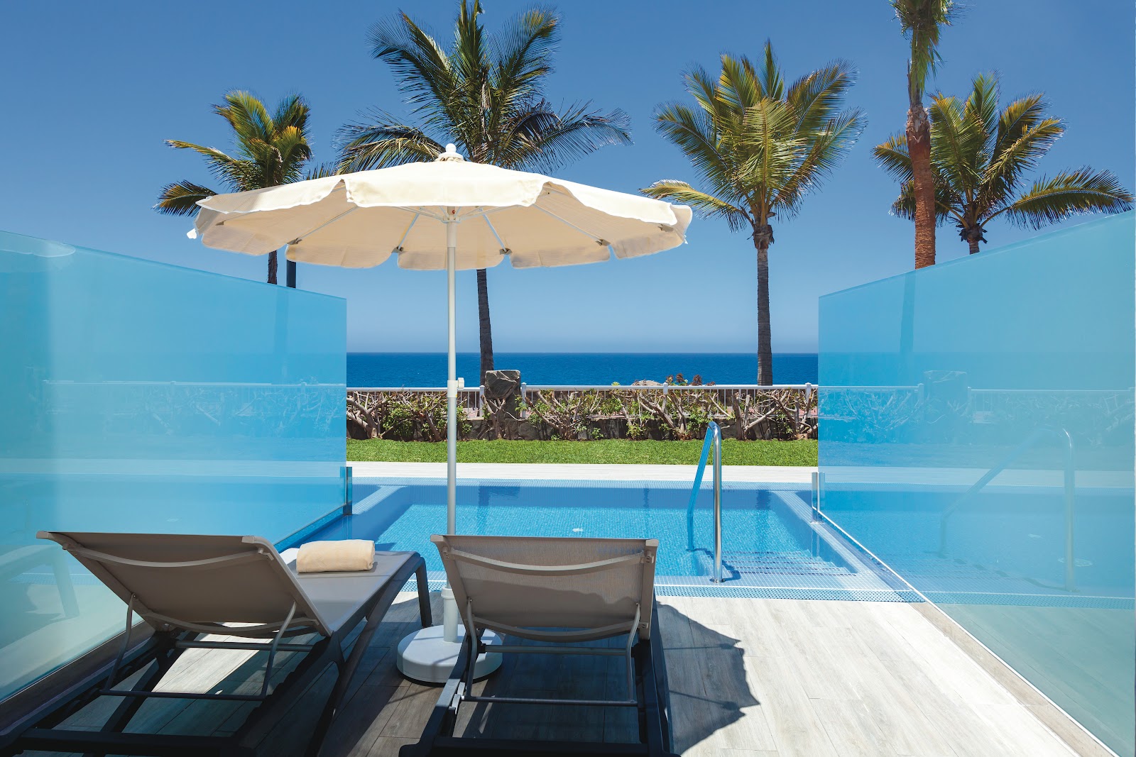 Two lounge chairs under a white umbrella overlook a pool with glass panels, palm trees, and the ocean beyond at Hotel Riu Gran Canaria.