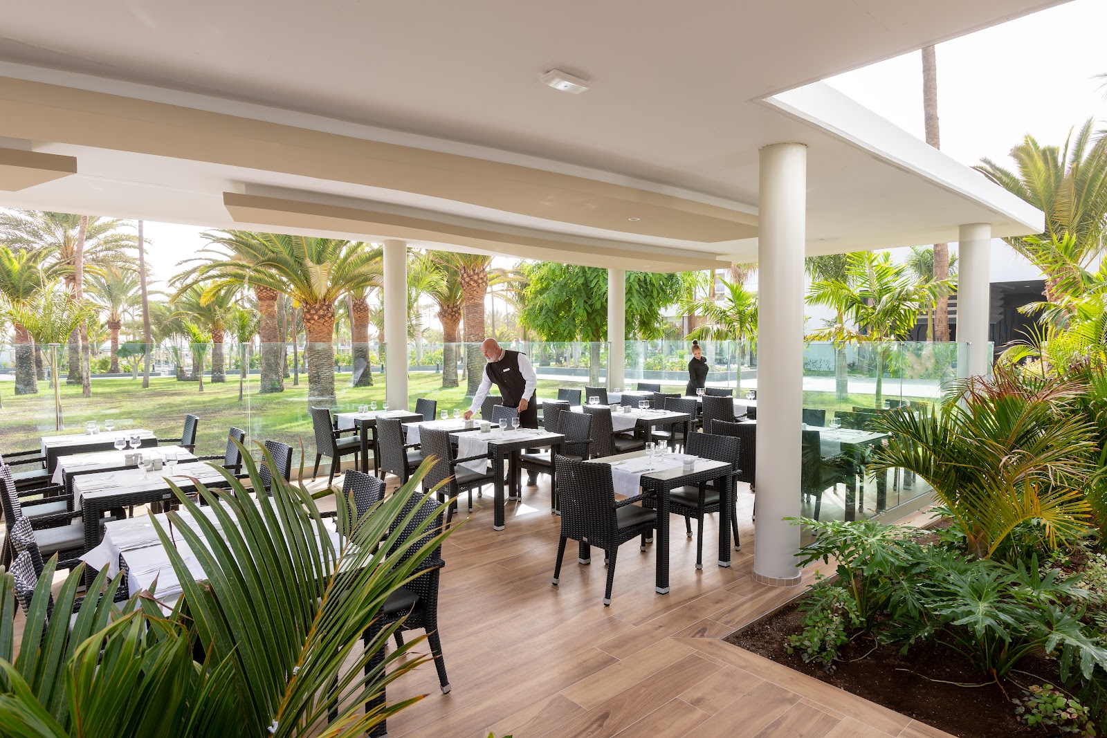 Open-air dining pavilion with black wicker chairs at white-topped tables, palm trees visible through glass walls and beyond the covered terrace.