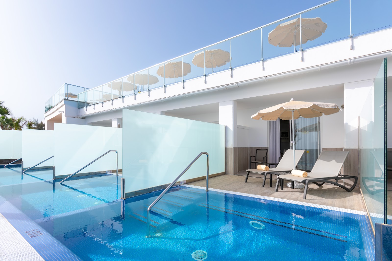 Modern resort suite with private pool access, lounge chairs, tan umbrellas, and glass barriers overlooking a larger blue pool below under clear sky.