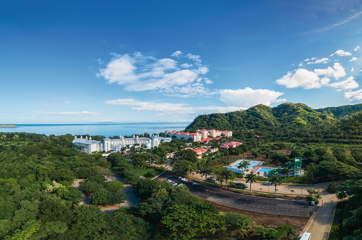 Resort buildings with red roofs and a blue water park nestled among dense green forests and hills overlooking a turquoise ocean under clear sky.