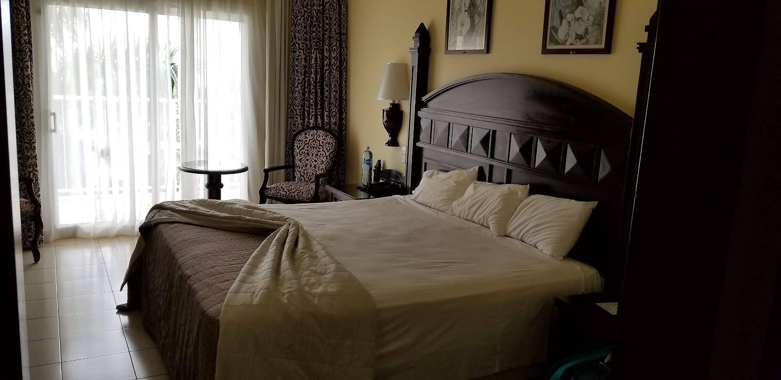 Hotel Riu Guanacaste guest room with queen bed, black headboard, yellow walls, patterned armchair, and glass doors opening to a balcony.