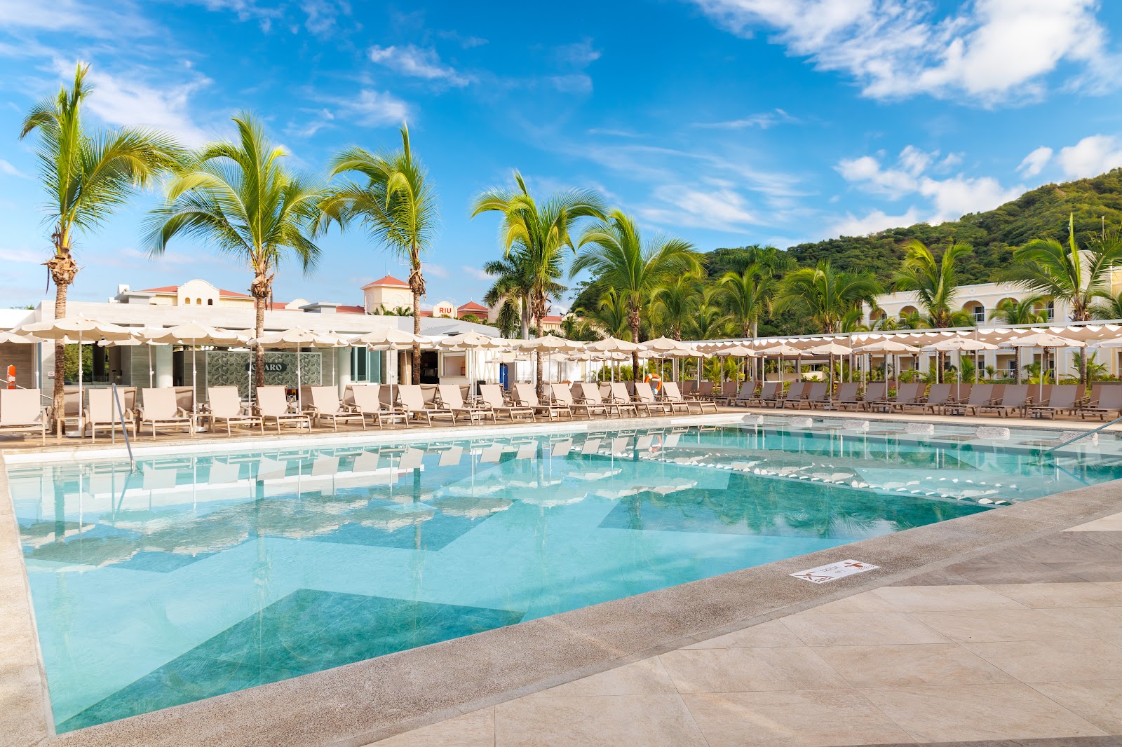 Turquoise pool with loungers and umbrellas lined along the deck, white resort buildings and palm trees in the background under blue sky.