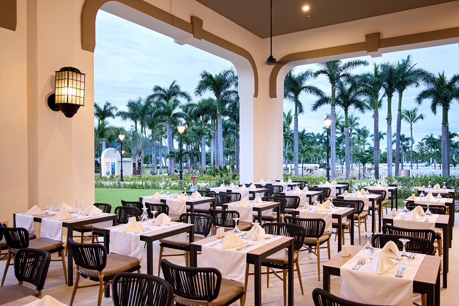Dining room with white-clothed tables and black woven chairs under an arched colonnade overlooking manicured grounds and towering palm trees.