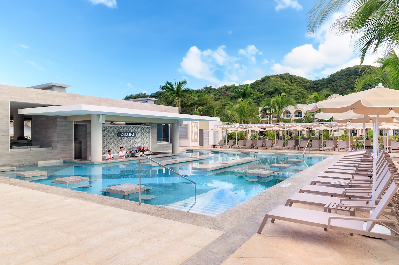 Resort swimming pool with lounge chairs, umbrellas, and palm trees under blue sky at Hotel Riu Guanacaste.