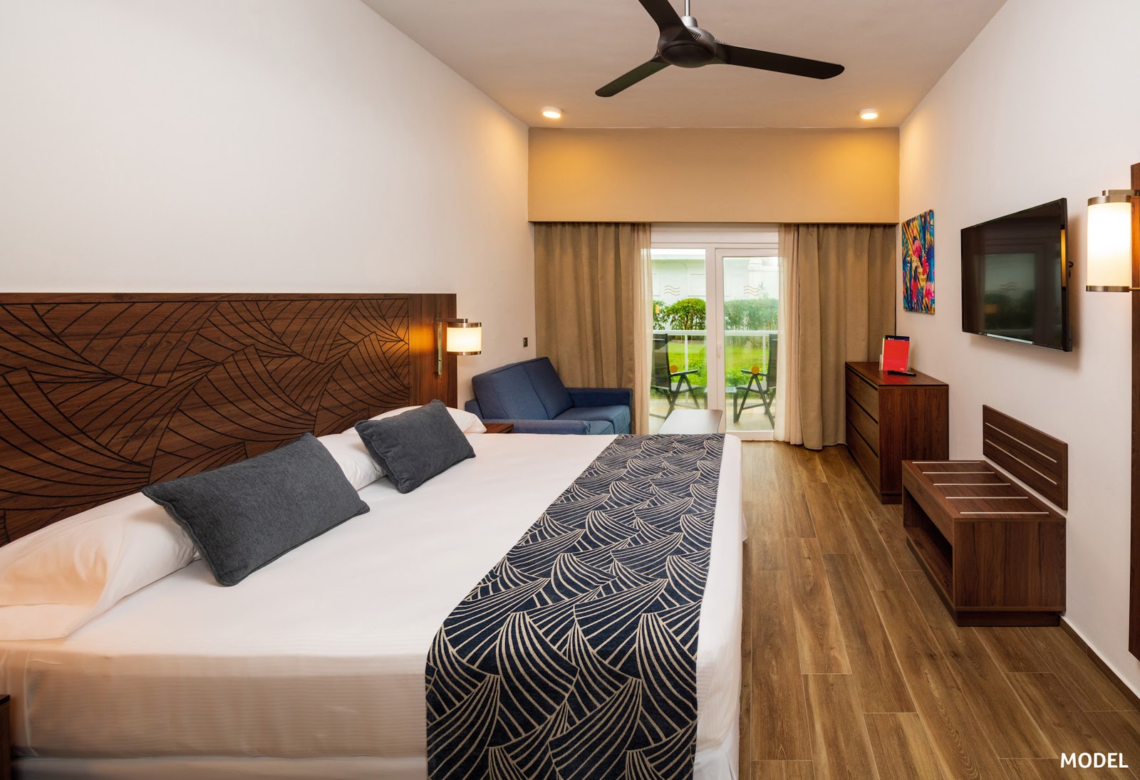 Resort guestroom with queen bed, wooden headboard, ceiling fan, wall-mounted TV, and glass door opening to balcony with lawn views.