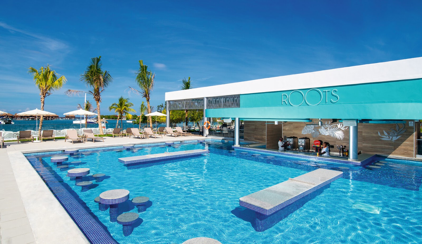 Large turquoise pool with sunken seating areas at Roots bar, palm trees, loungers with umbrellas, and ocean view beyond.