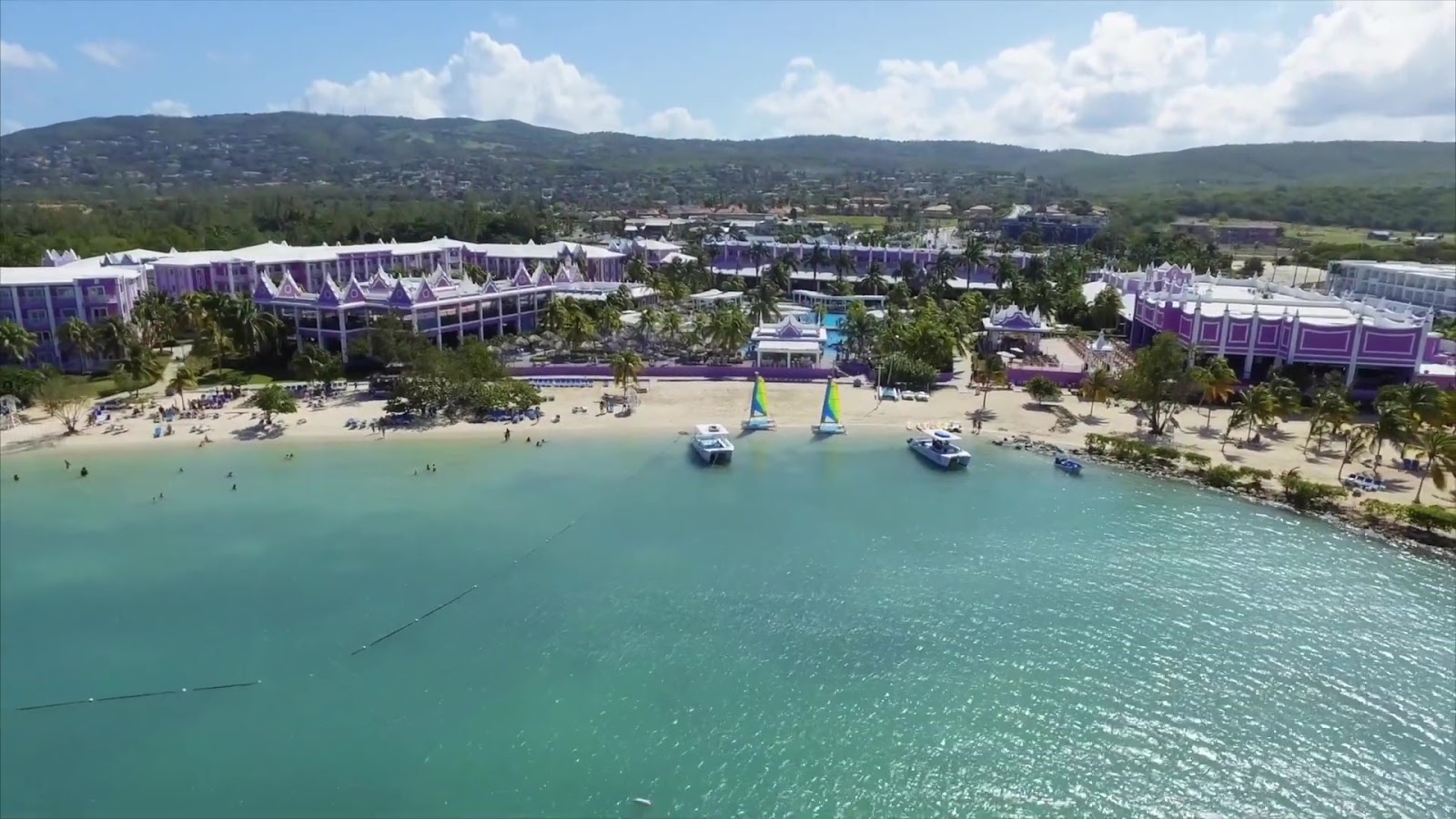 Aerial view of Hotel Riu Montego Bay's purple buildings, beach with swimmers, and turquoise water with sailing boats offshore.
