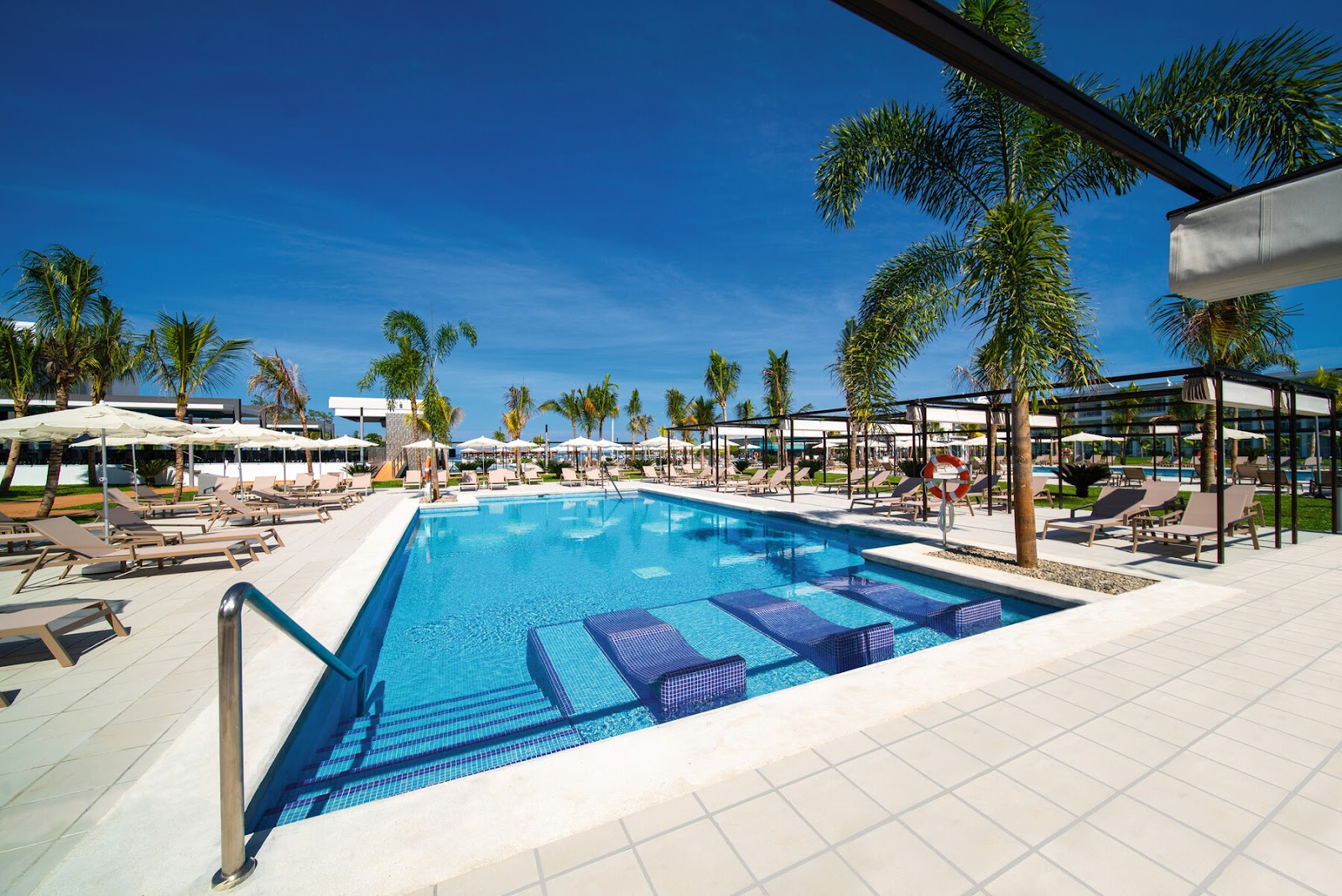 Resort pool with submerged loungers, surrounded by palm trees, white umbrellas, and cabanas under clear blue sky.