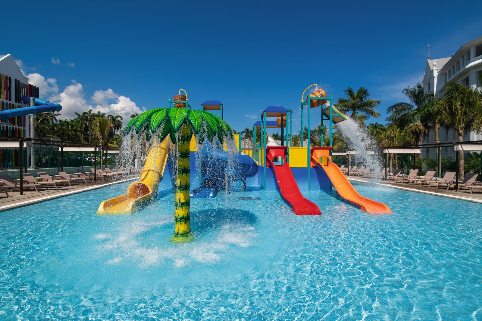Colorful water slides with yellow, red, and orange tubes cascade into a shallow pool at Hotel Riu Ocho Rios, surrounded by palm trees and lounge chairs.