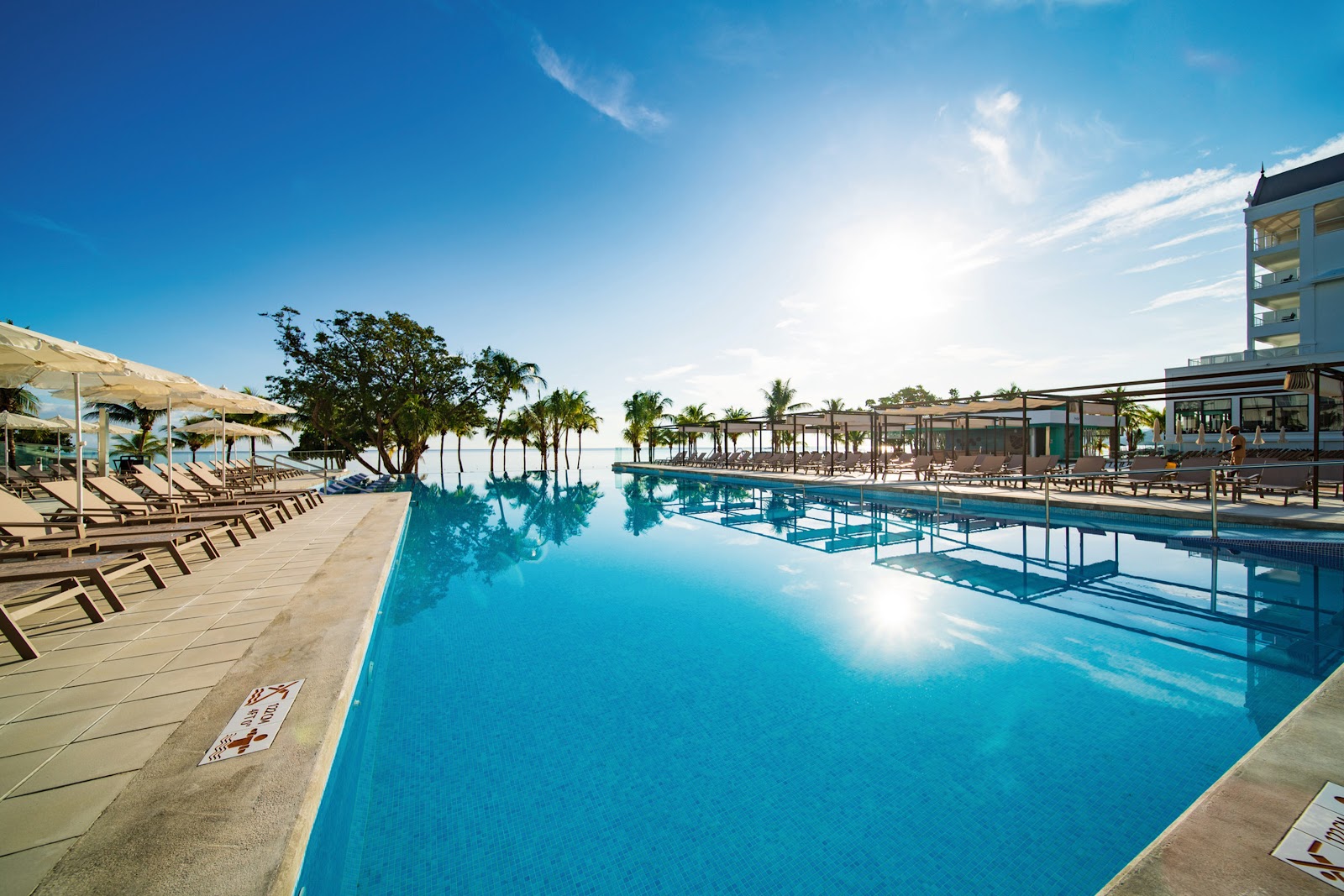 Large swimming pool with lounge chairs and umbrellas, palm trees, and a multi-story hotel building under clear blue sky.