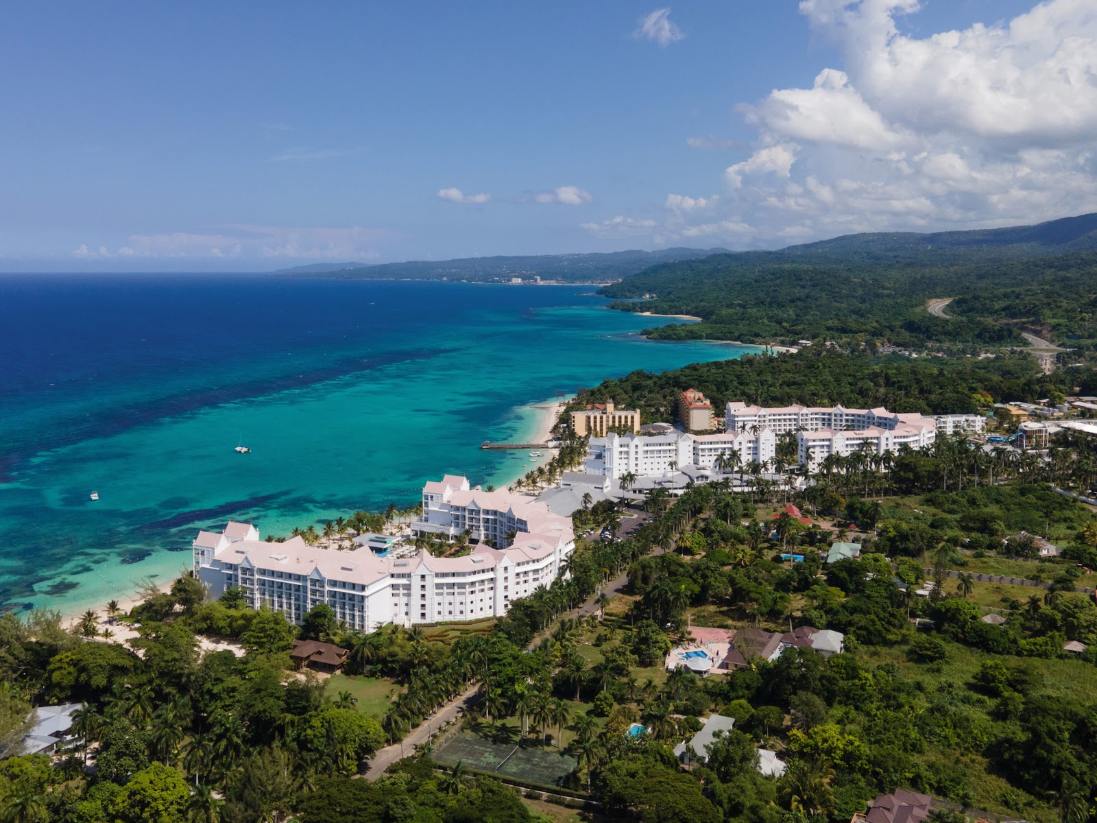 White resort buildings curve along a sandy beach bordered by turquoise Caribbean waters and lush tropical forest in Jamaica.