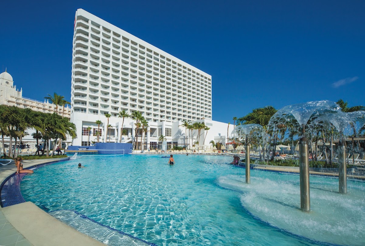 Large white high-rise hotel tower rises behind a sprawling turquoise pool with swim-up bar, surrounded by palm trees and lounge chairs under clear blue sky.