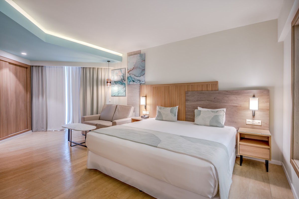 Modern resort bedroom with king bed, wooden nightstands, sitting area, and turquoise ceiling accent lighting at Hotel Riu Palace Antillas.
