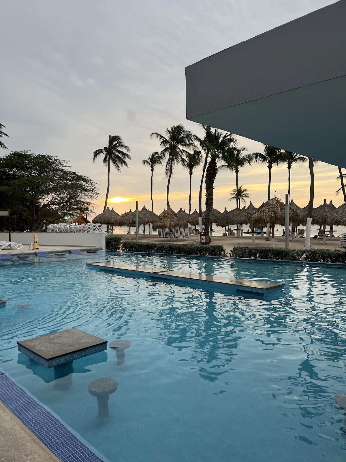 Resort pool with submerged loungers at sunset, palm trees and thatched umbrellas beyond, modern building overhead.